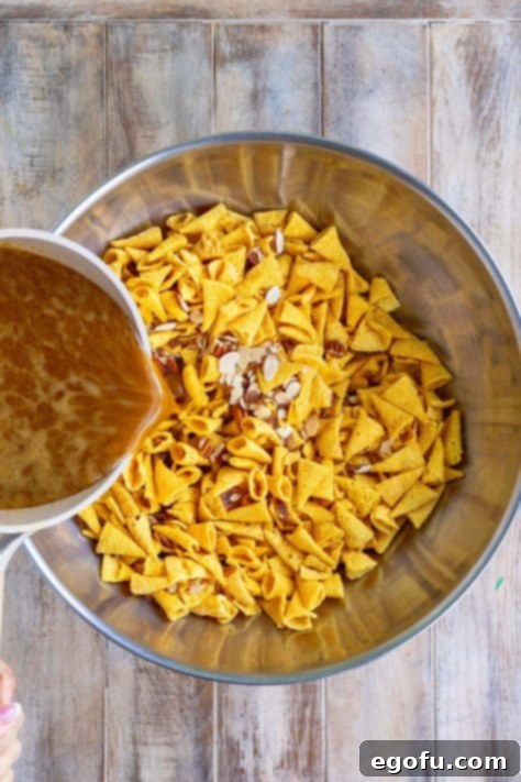 Sweet sauce being poured over the party mix ingredients, ensuring an even coating.