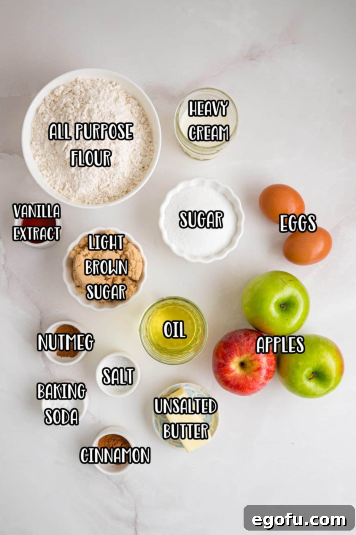 A flat lay photograph of all-purpose flour, heavy cream, vanilla extract, granulated sugar, light brown sugar, eggs, vegetable oil, diced apples, ground nutmeg, salt, baking soda, unsalted butter, and ground cinnamon, neatly arranged as ingredients for Apple Dapple Cake.