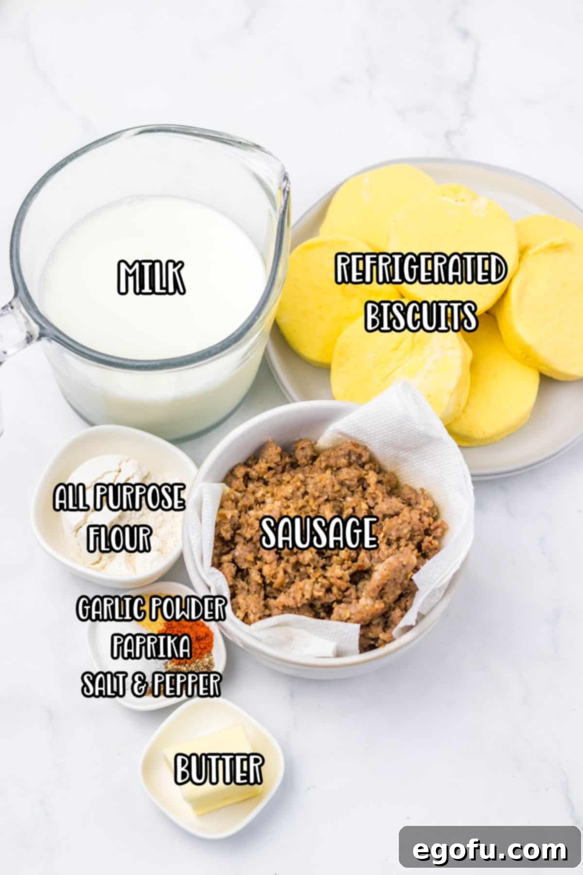 A vibrant flat lay of all the essential ingredients: milk, refrigerated biscuits, sausage, flour, butter, salt, pepper, black pepper, garlic powder, paprika, and fresh parsley.