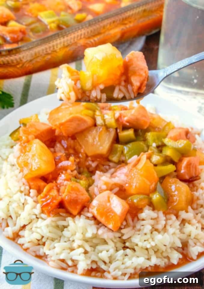 Sticky Sweet-Sour Chicken Bake 7 A forkful of baked sweet and sour chicken and rice, showcasing the tender chicken, pineapple, and peppers, with a full serving on a plate below.