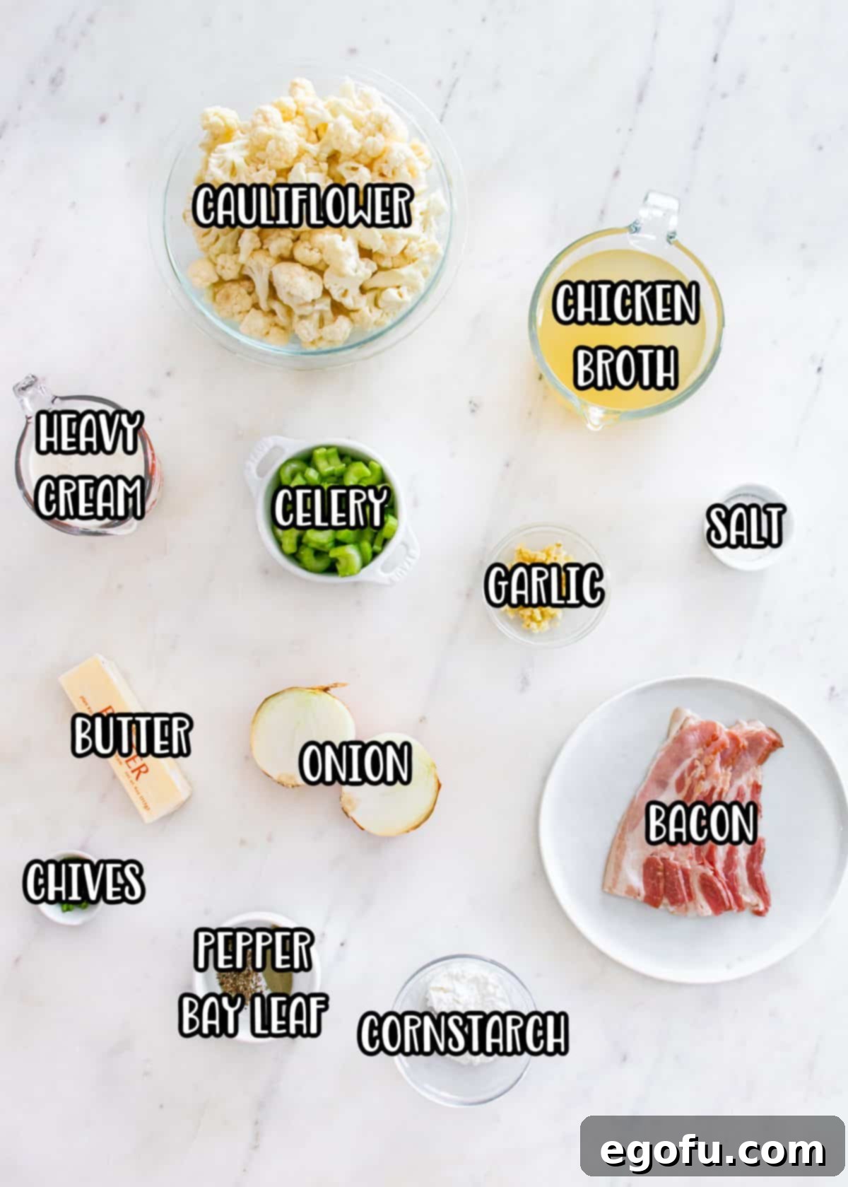 Various ingredients for Loaded Cauliflower Soup laid out: bacon, butter, yellow onion, celery, salt, black pepper, garlic, cauliflower, chicken broth, bay leaf, heavy whipping cream, cornstarch, cold water, and fresh chopped chives.