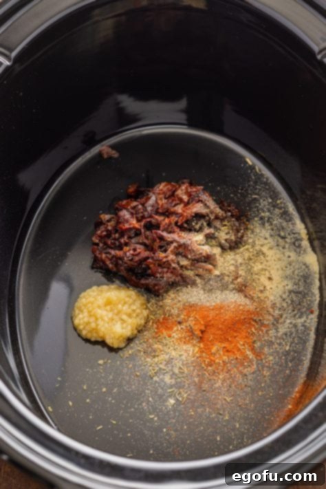 A Crock Pot with minced garlic, sun dried tomatoes, tomato paste, onion powder, Italian seasoning, paprika and black pepper.