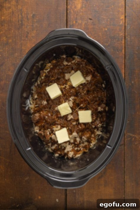 Slabs of butter on onion, seasonings, and beef in a Slow Cooker.