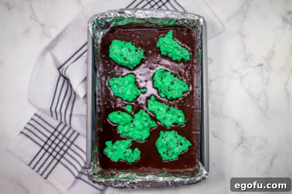 brownie batter in a baking pan with bright green cream cheese dotted on top of the brownie batter