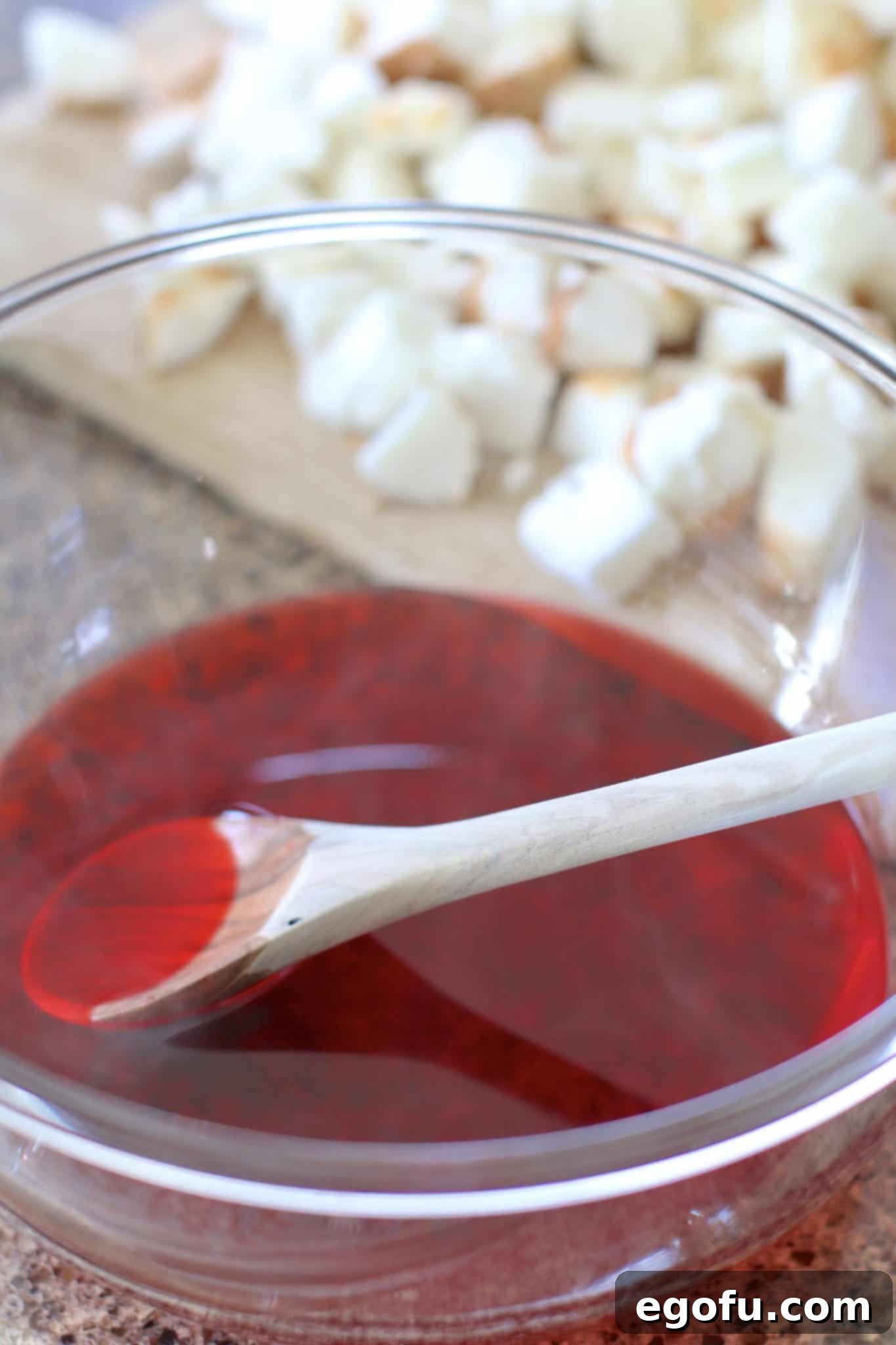 Strawberry Angel Delight 7 Strawberry gelatin powder being mixed with hot water in a clear glass bowl using a wooden spoon, creating a vibrant red solution.