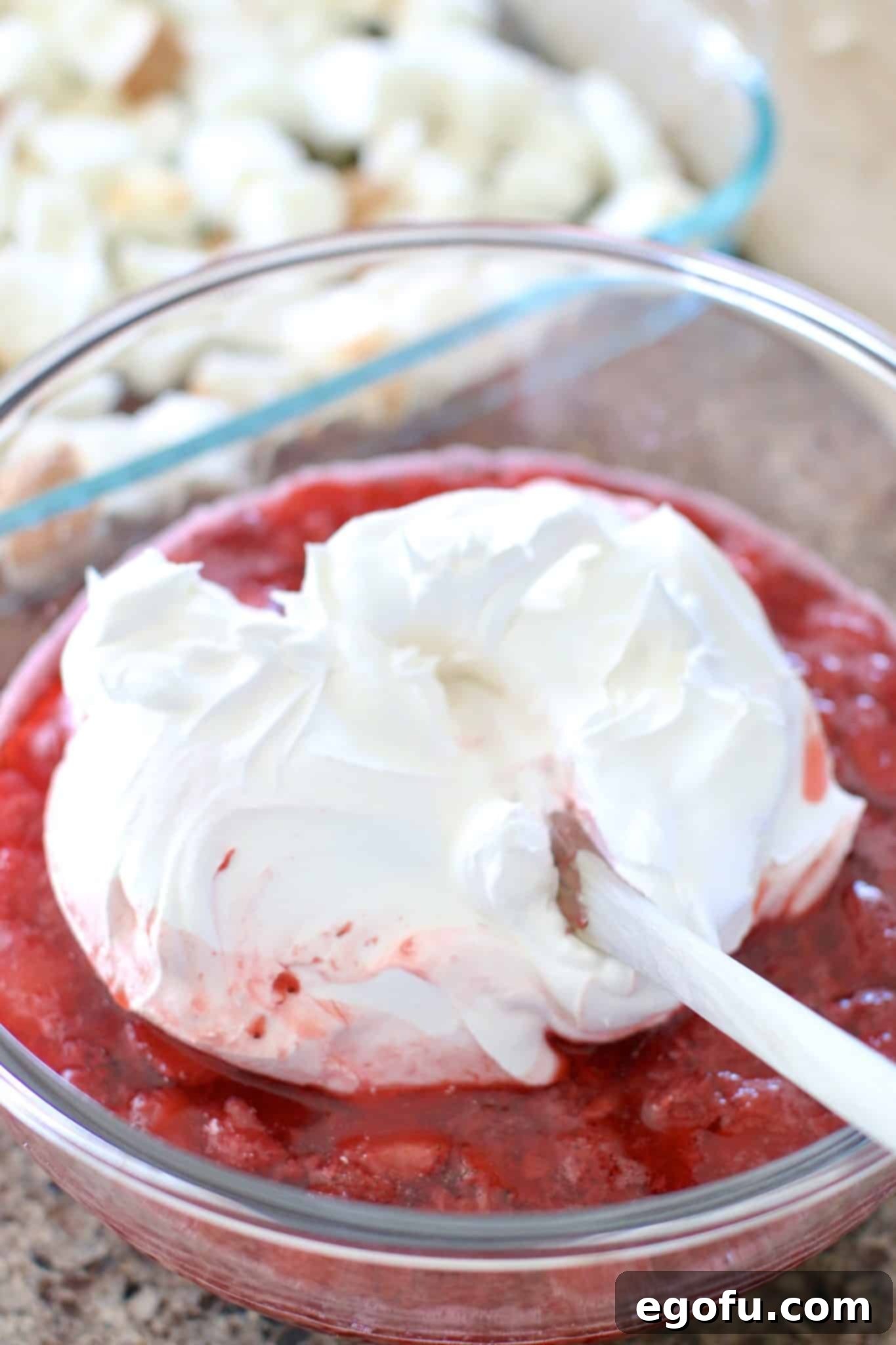Strawberry Angel Delight 9 Creamy Cool Whip being gently folded into the strawberry and gelatin mixture in a clear glass bowl with a wooden spoon, creating a light pink swirl.
