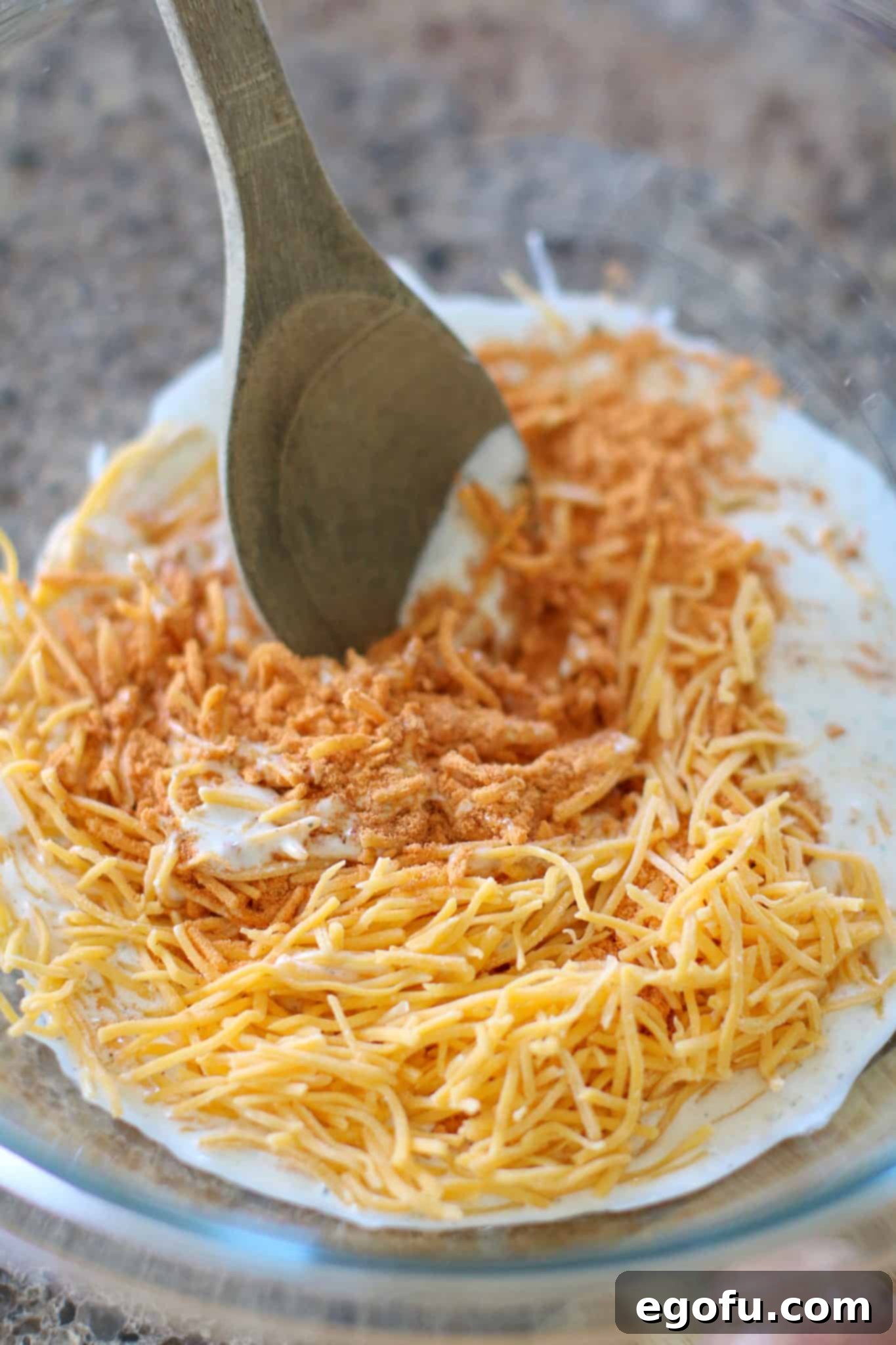 a wooden spoon in a clear bowl with taco seasoning, ranch dressing and shredded cheese.