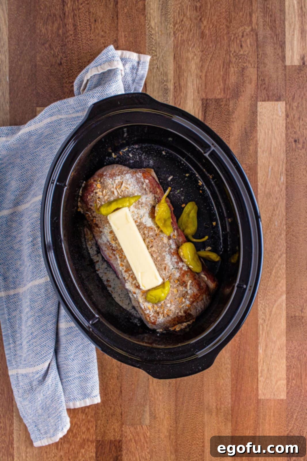 Fall-Apart Mississippi Pork Perfection 9 A stick of butter placed on a pork roast with peperoncini peppers around it in an oval slow cooker.