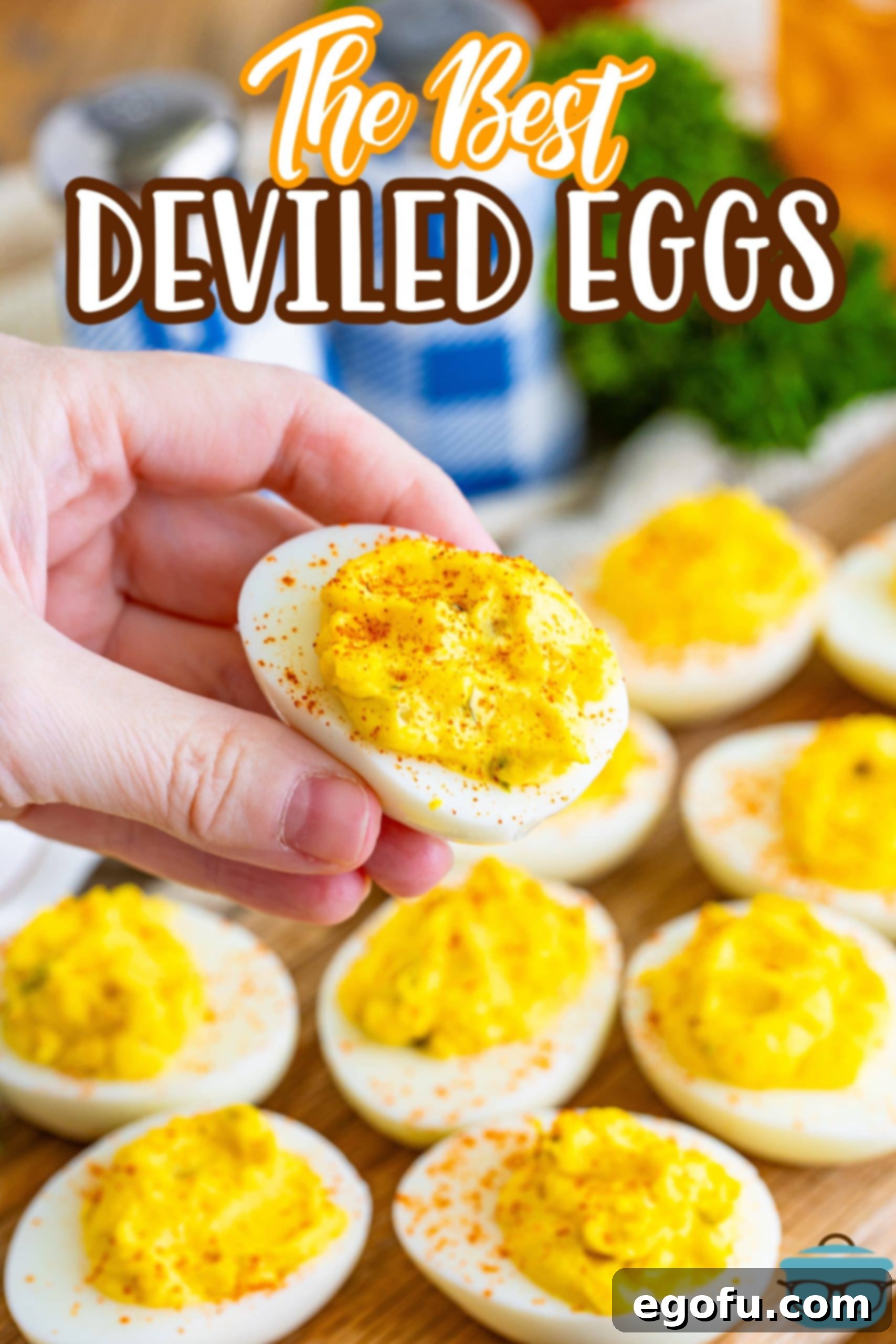 A hand holds up a single perfectly garnished deviled egg over a platter of other deviled eggs on a wooden serving board.
