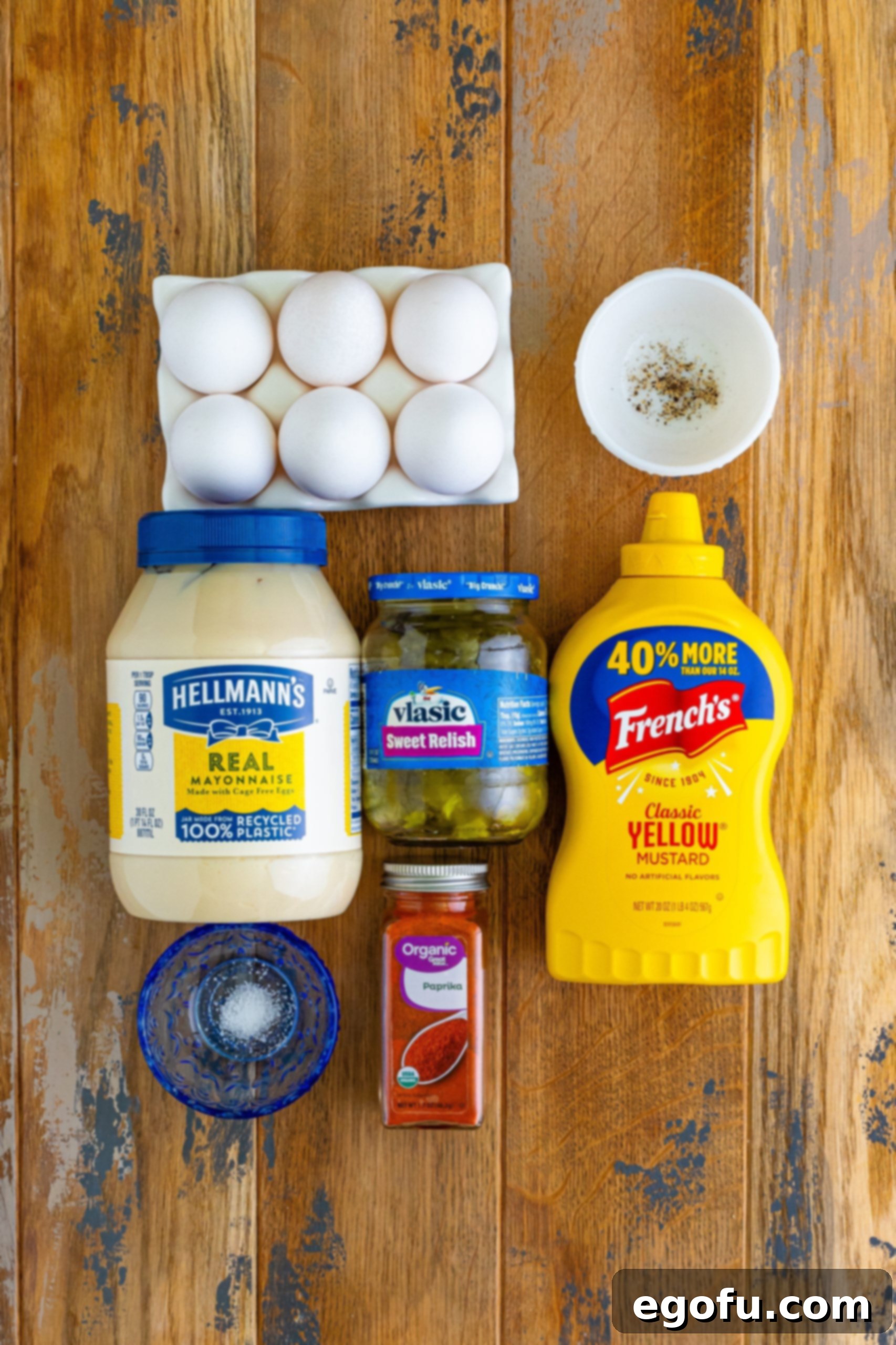 A flat lay photograph showcasing the key ingredients for deviled eggs: 6 large eggs, mayonnaise, yellow mustard, sweet pickle relish, salt, pepper, and paprika.