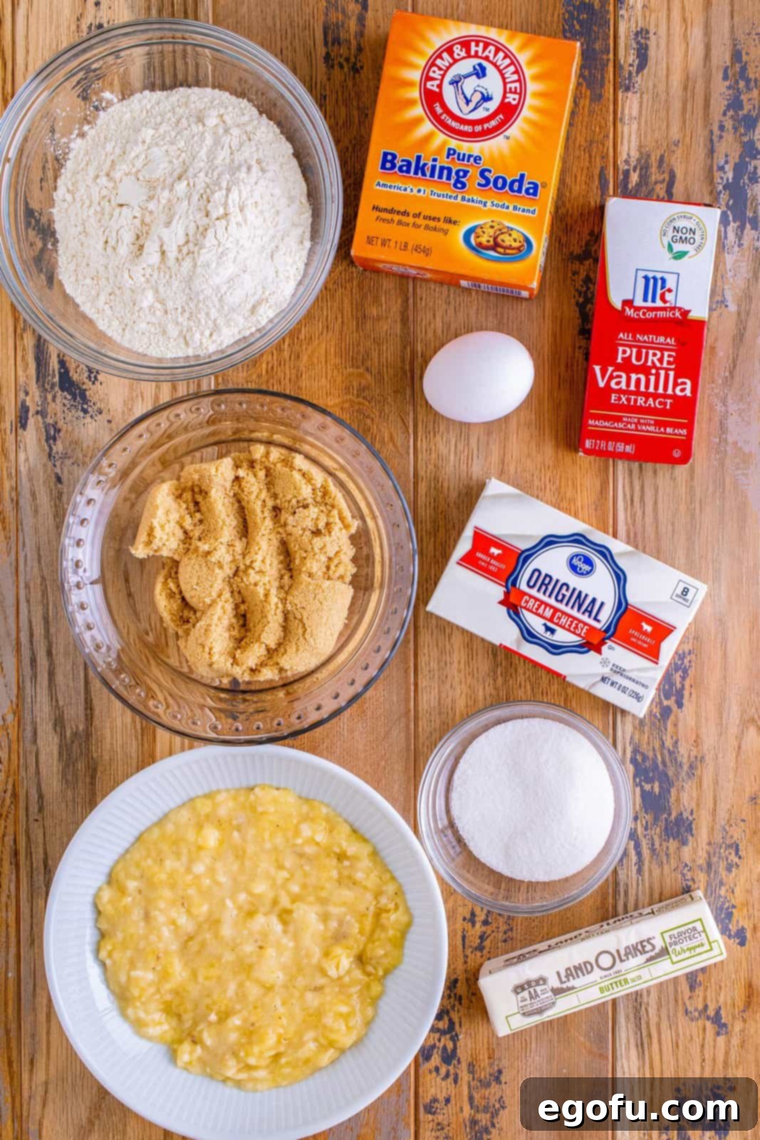 Cream Cheese Swirl Banana Bread 4 An array of ingredients for cream cheese banana bread, including bananas, butter, brown sugar, egg, vanilla extract, baking soda, flour, cream cheese, and sugar, neatly arranged for baking.