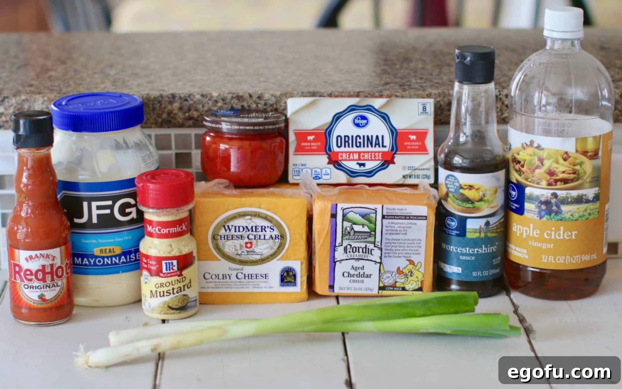 A vibrant flat lay of all ingredients laid out for homemade pimento cheese: cream cheese, mayonnaise, Worcestershire sauce, apple cider vinegar, hot sauce, sugar, ground mustard, a jar of diced pimiento, blocks of Colby Cheese and Aged Cheddar Cheese, and green onions.