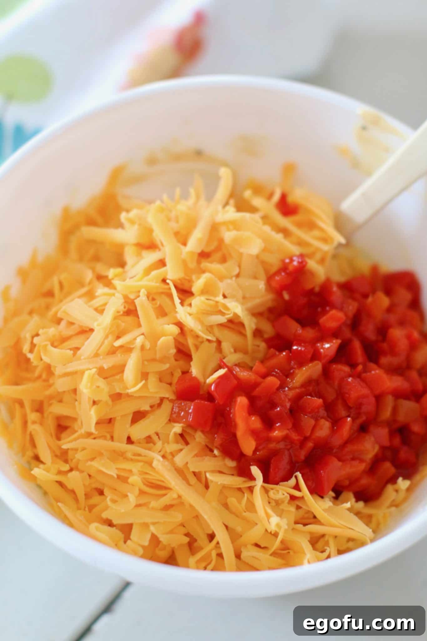 Shredded cheddar cheese and diced pimentos being folded into the creamy cream cheese mixture in a white bowl, showing the blend of ingredients.