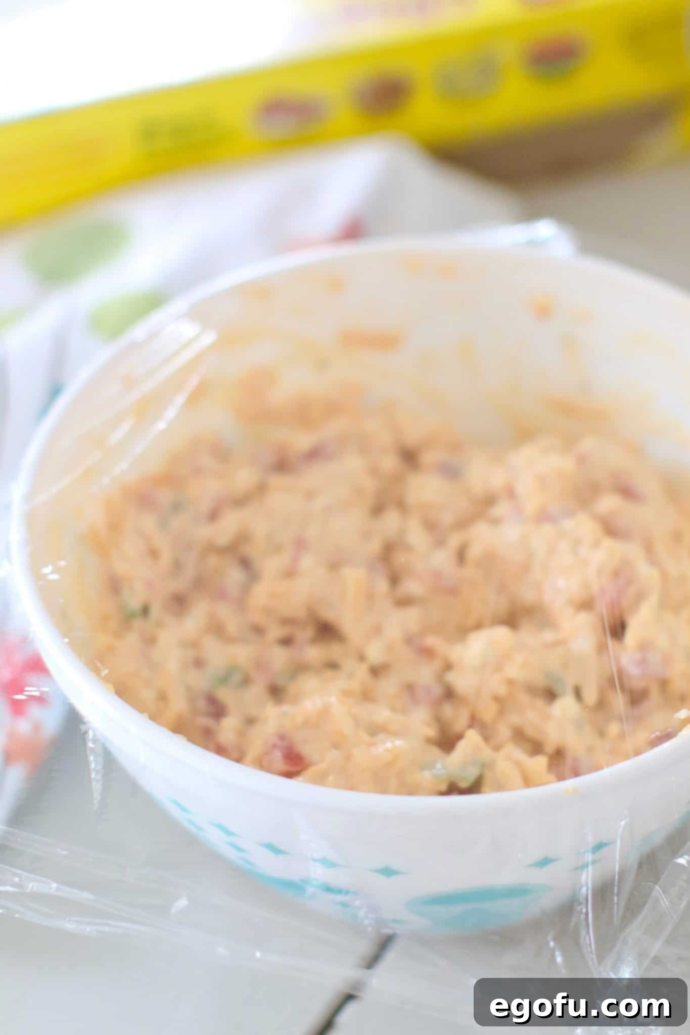 A bowl of freshly mixed pimento cheese spread, covered tightly with plastic wrap and placed in the refrigerator to chill and allow flavors to meld.