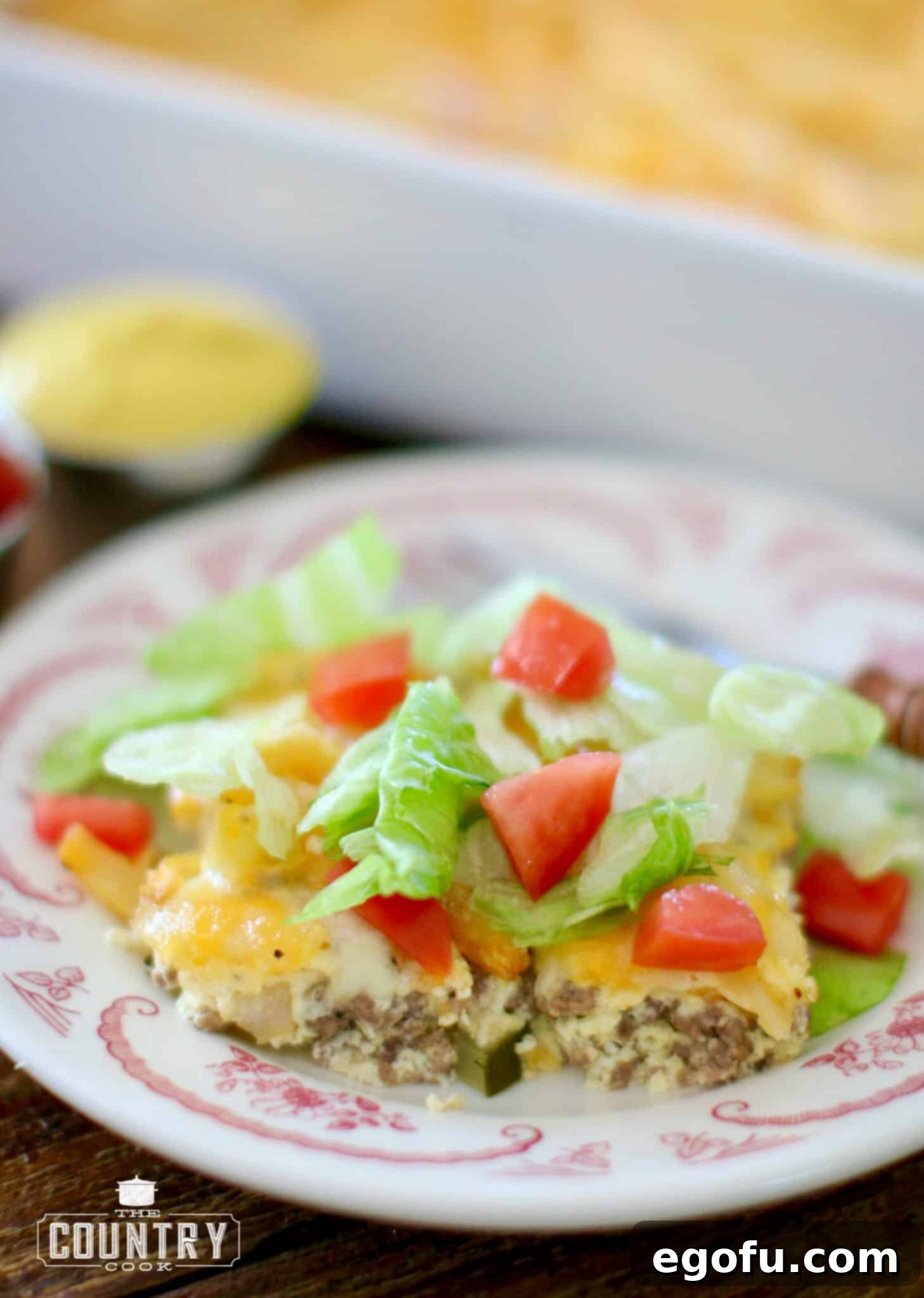 A slice of cheeseburger and fries casserole served on a plate and topped with shredded lettuce and diced tomatoes.