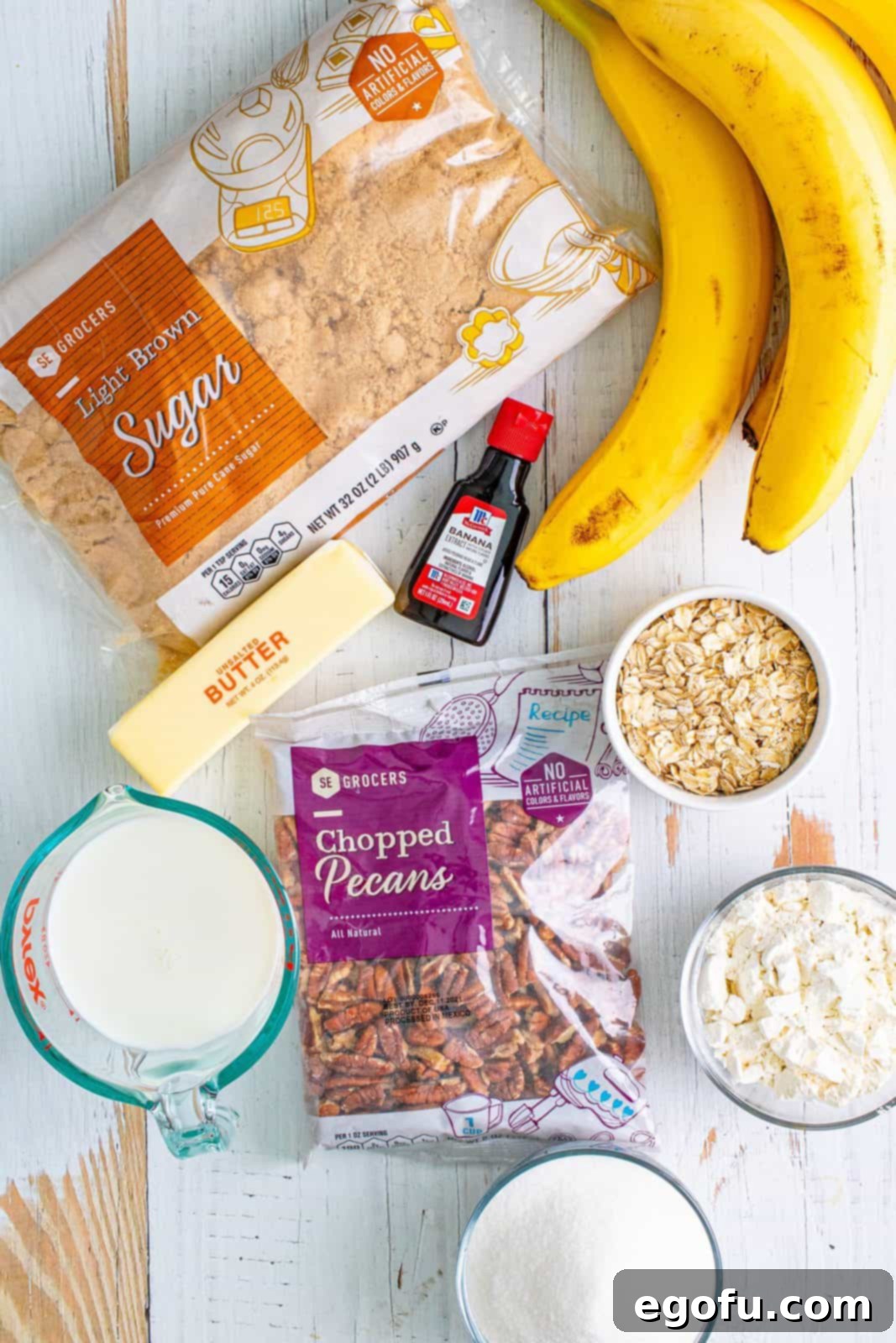 An array of fresh ingredients neatly arranged on a countertop, including a bowl of self-rising flour, a stick of unsalted butter, uncooked old-fashioned oats, a pile of chopped pecans, granulated sugar, a glass of milk, and several ripe bananas, ready for baking this delicious cobbler.