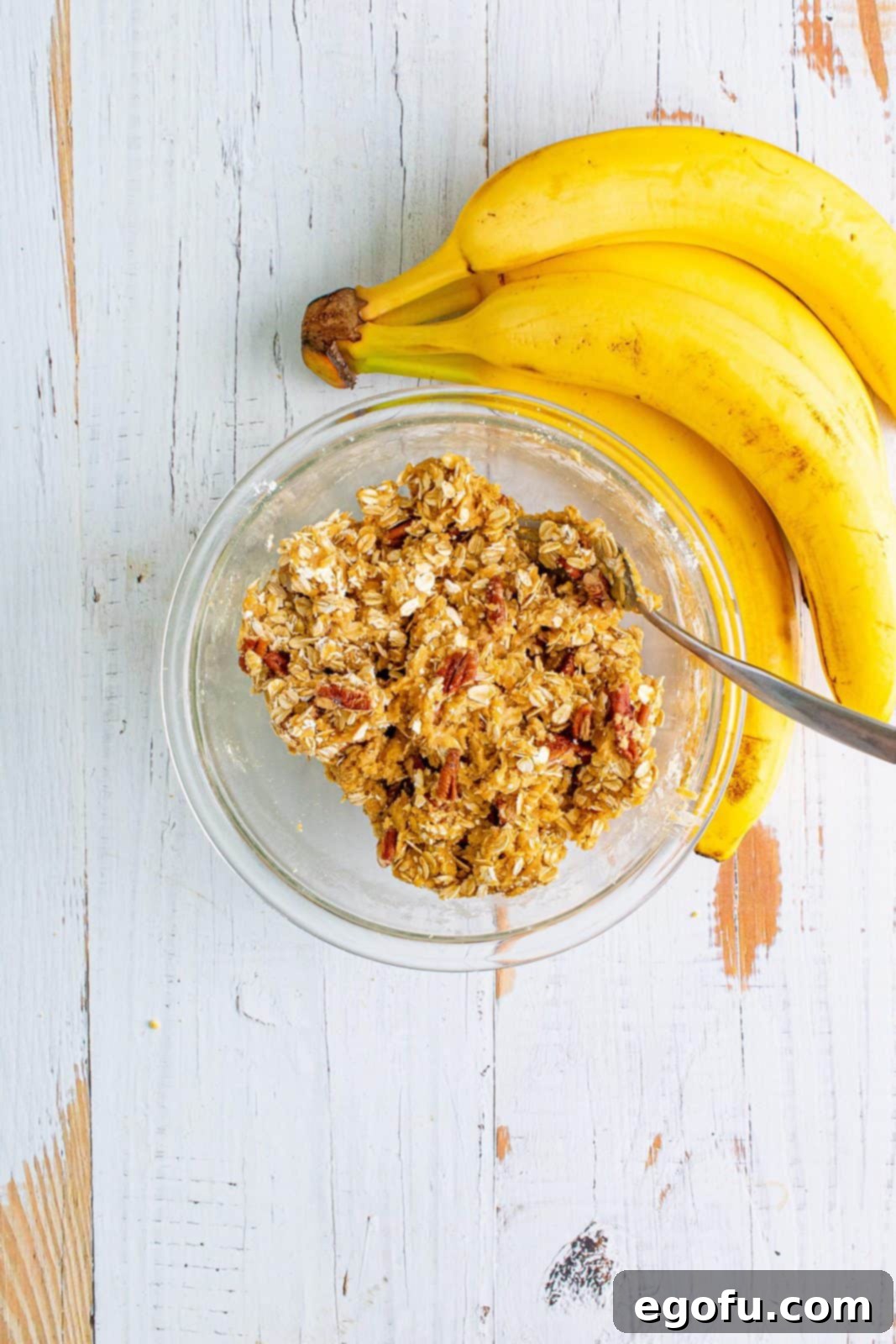 A glass bowl holds a rich brown mixture of brown sugar, self-rising flour, and softened butter, mixed until crumbly. Uncooked old-fashioned oats and chopped pecans are visible, awaiting final incorporation to form the streusel topping.