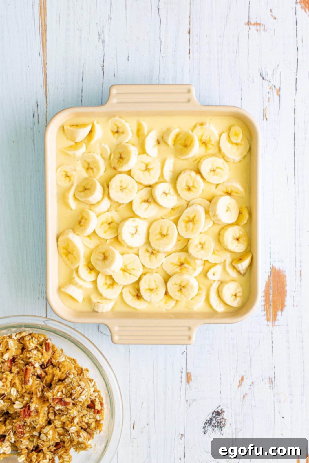 Neatly sliced ripe bananas are layered meticulously on top of the creamy cobbler batter in a square baking pan, preparing for the streusel topping.