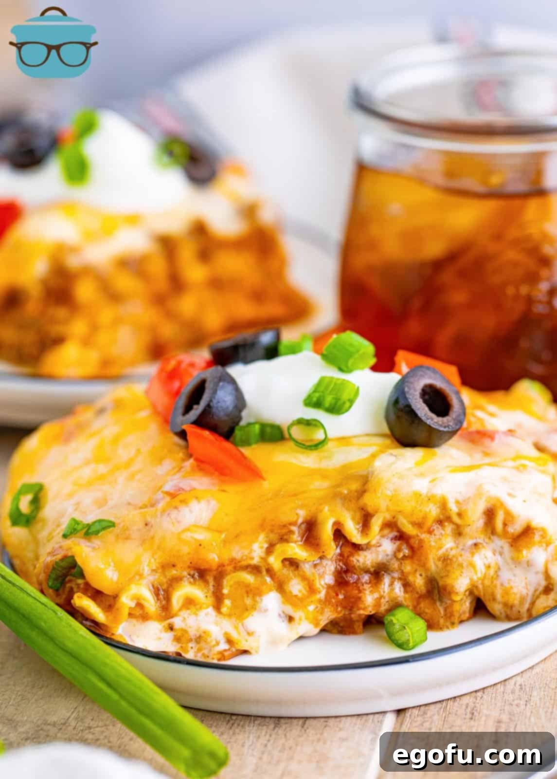 A slice of Mexican Lasagna topped with sour cream and green onions on a white plate, with fresh limes and cilantro in the background.