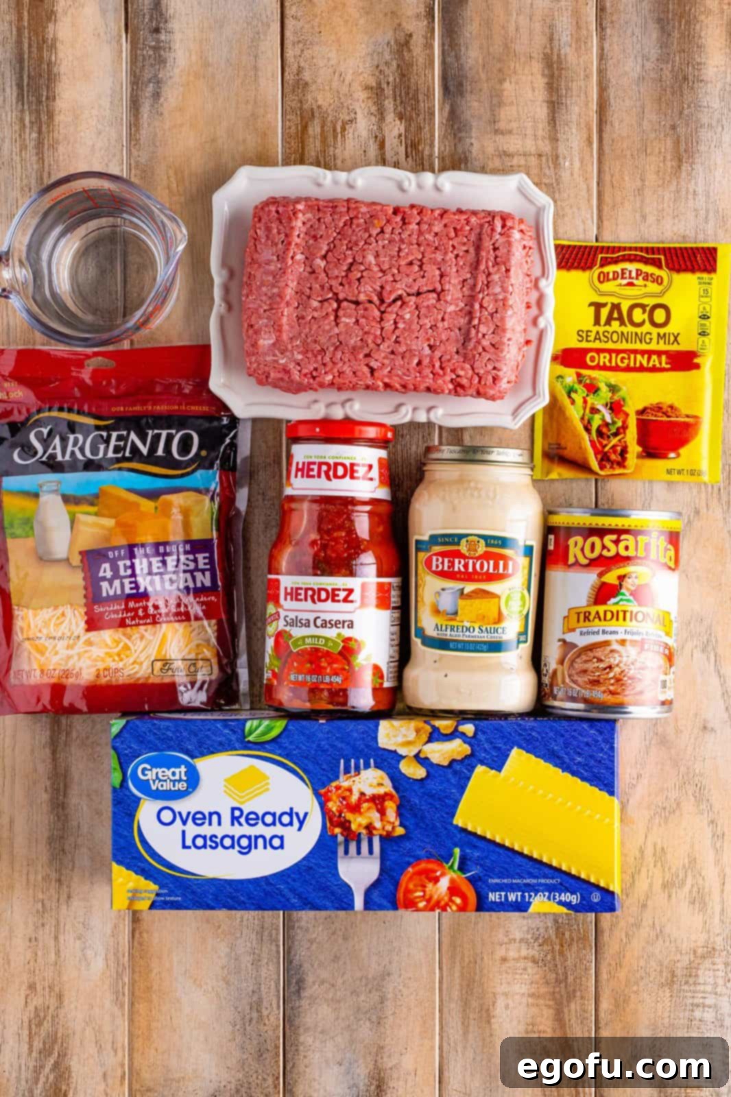 A vibrant flat lay of all the fresh ingredients needed for Mexican Lasagna: ground beef, taco seasoning, Alfredo sauce, salsa, oven-ready lasagna noodles, refried beans, water, and shredded cheese.