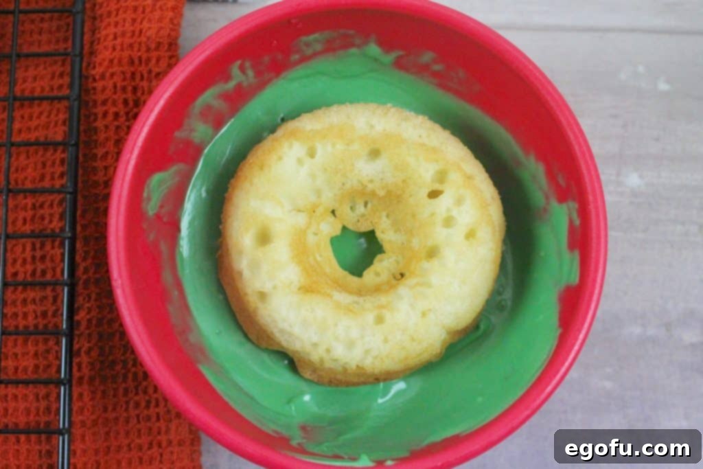 A baked doughnut being dipped into a bowl of melted green candy melts.