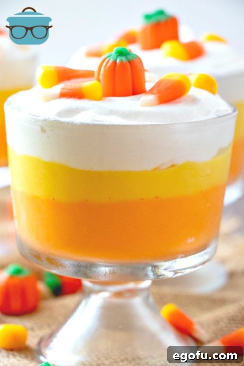 Vibrant Candy Corn colored layered vanilla pudding in elegant glass dessert cups, beautifully topped with fluffy whipped cream, real candy corn pieces, and adorable mallow creme pumpkins, ready for a festive Halloween celebration.