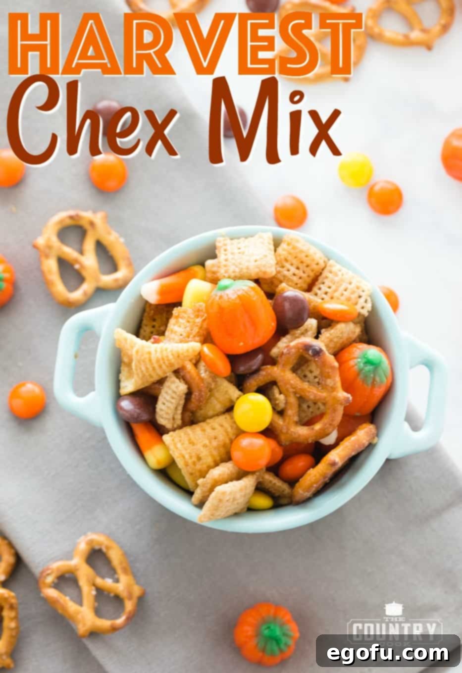 Harvest Chex Mix recipe from The Country Cook. Chex mix shown in a small white bowl with pretzels and Reese's pieces candy scattered around it, highlighting the vibrant colors and delightful ingredients.