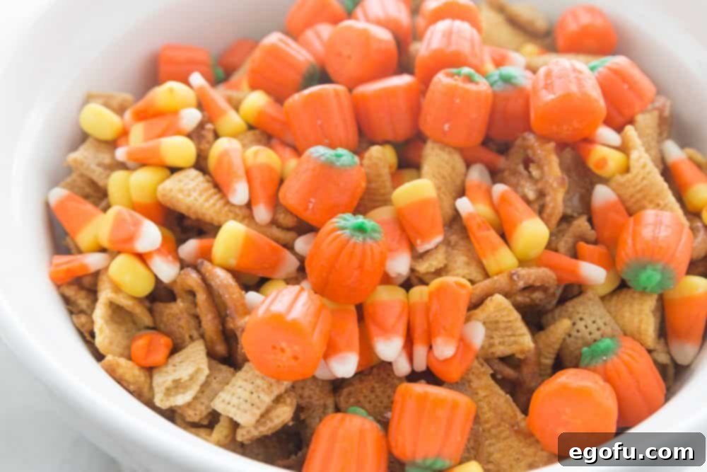Candy corn and mallow creme pumpkins shown on top of cooled chex mix in a white bowl, ready for the final toss.