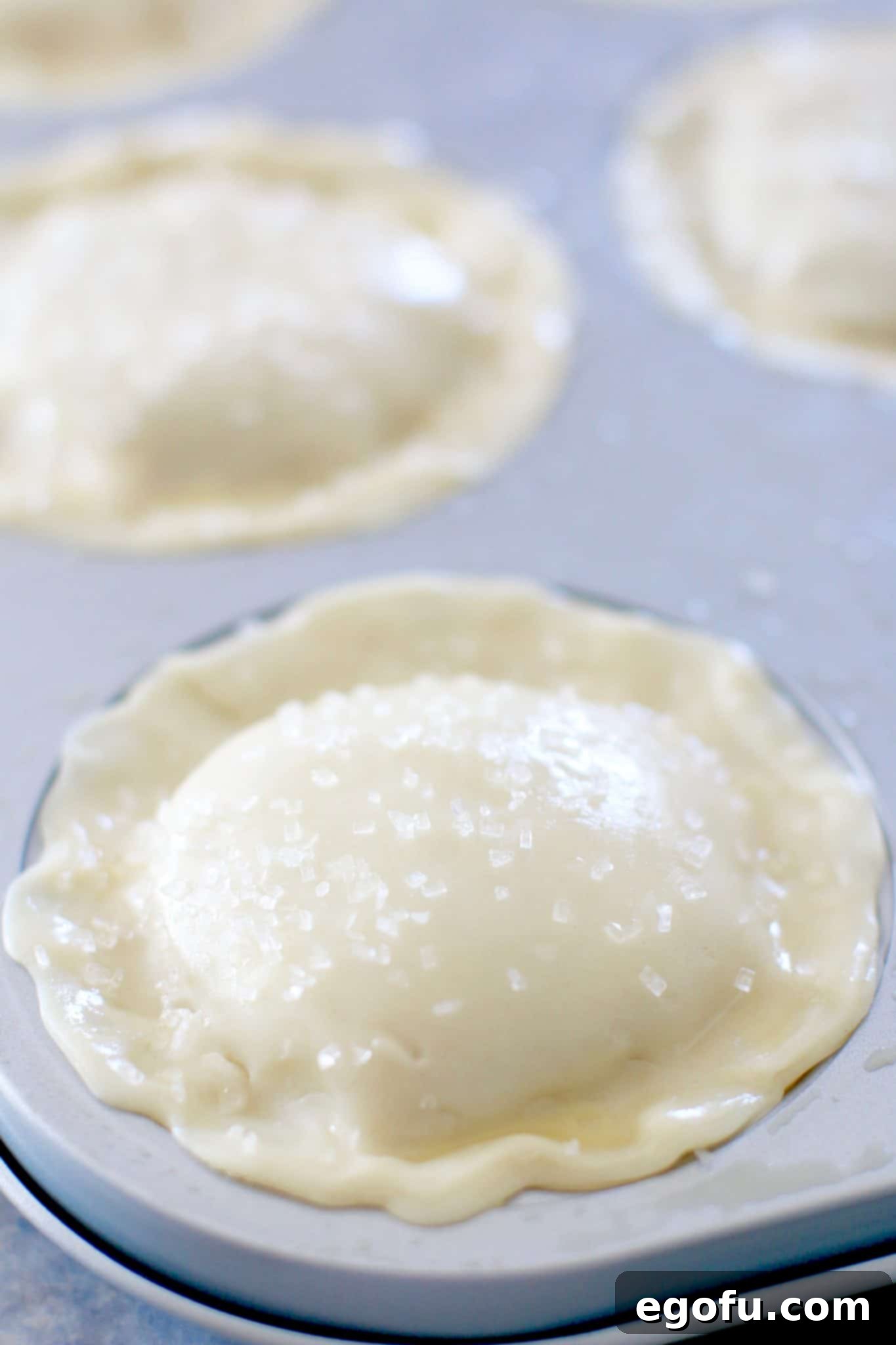 Cherry Cheesecake Minis 15 Mini cherry pies, freshly brushed with egg wash and sprinkled with sugar, arranged on a baking sheet, ready for the oven.