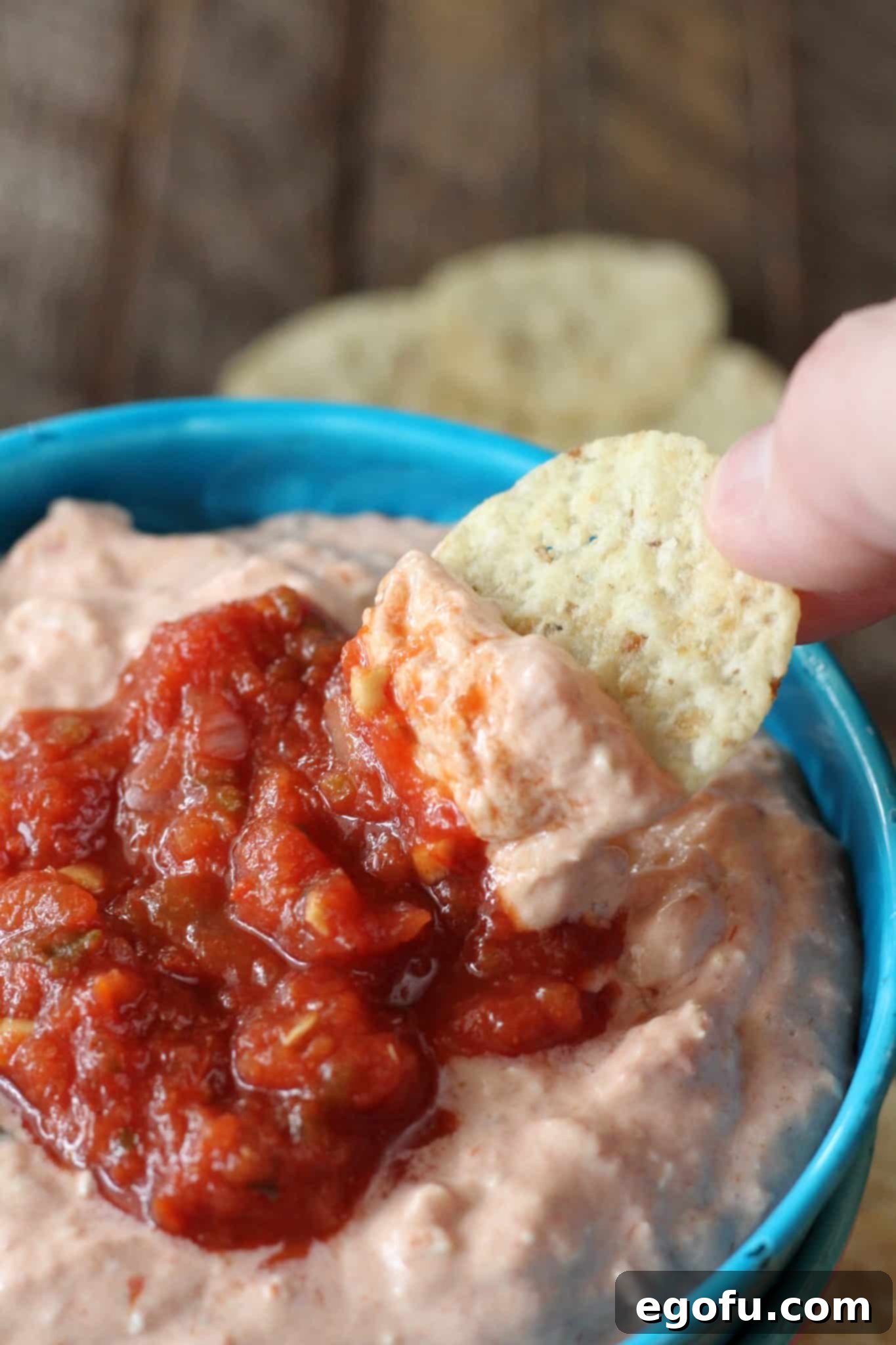a hand shown dipping a tortilla chip into the chicken dip in the blue bowl.