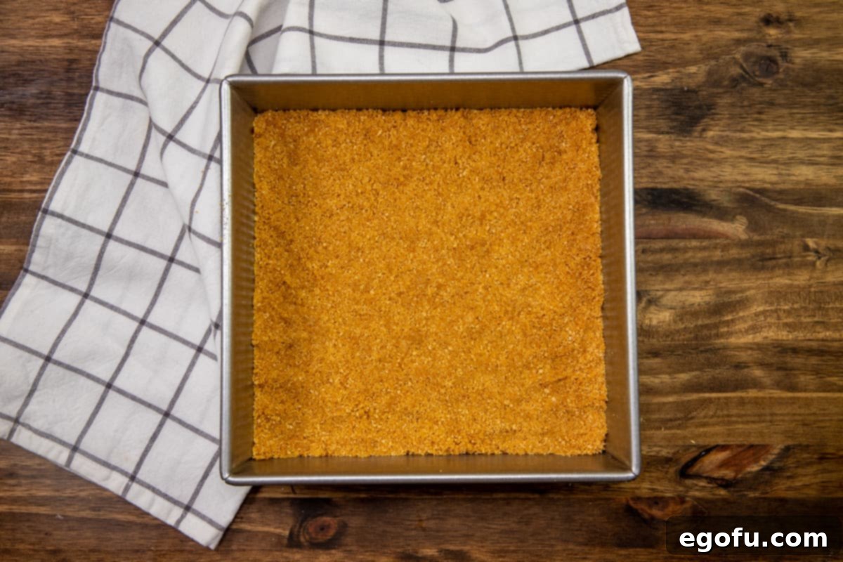 The graham cracker crust mixture spread and pressed into the bottom of a 9x9 inch metal baking pan, forming a flat, even layer.