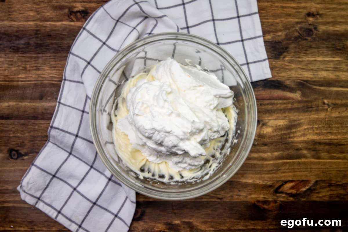 Freshly made homemade whipped cream being poured over the cream cheese mixture in a bowl, before being gently folded together.