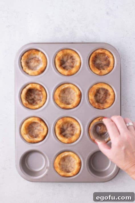 A hand using a shot glass to shape the warm biscuit dough into deeper cups in a muffin tin.