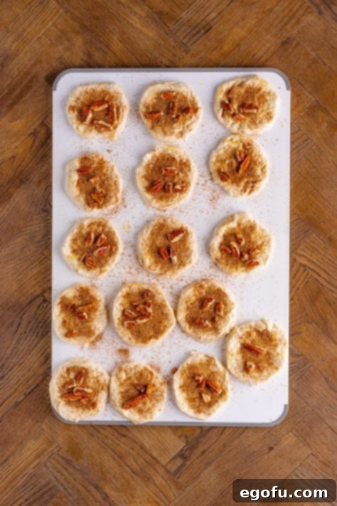 Cinnamon-pecan mixture being sprinkled on top of buttered raw biscuit slices.
