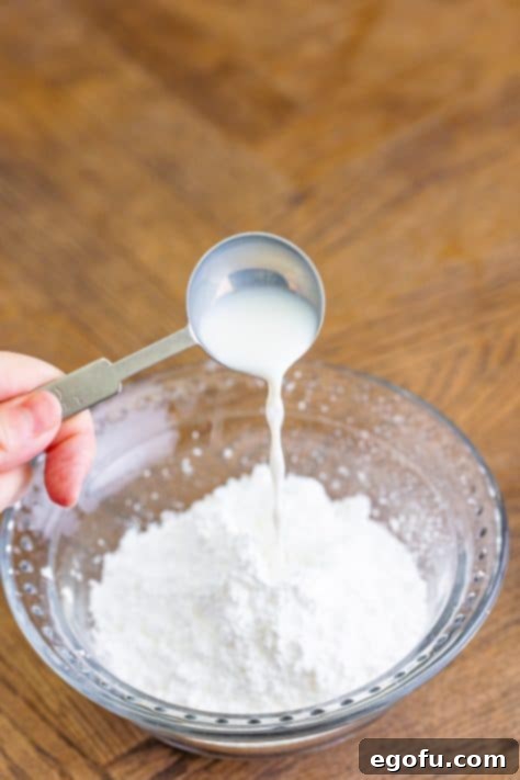 A measuring spoon pouring milk into powdered sugar in a bowl to make icing.