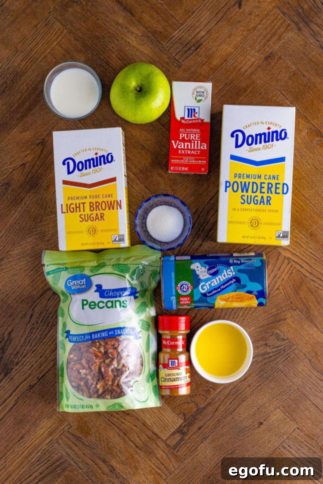 A vibrant flat lay of all ingredients for Cinnamon Apple Pull-Apart Bread: green apple, brown sugar, powdered sugar, chopped pecans, canned biscuits, cinnamon sticks, butter, milk, and granulated sugar.