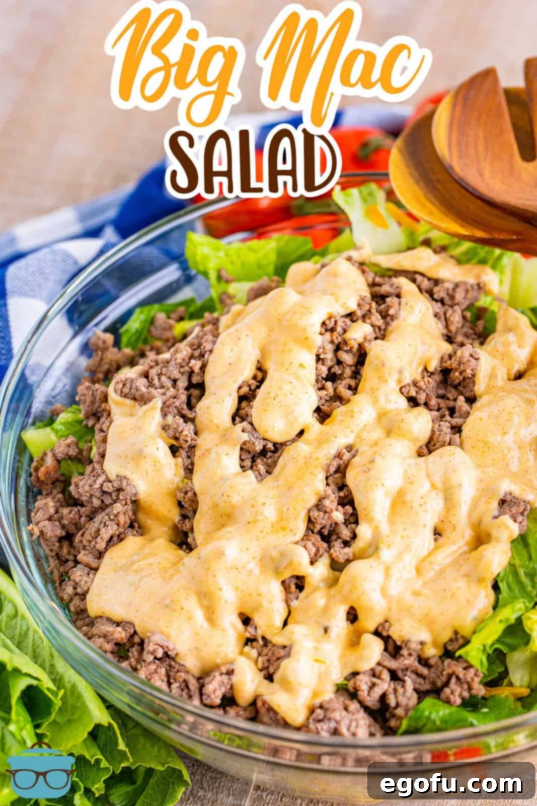 A vibrant Big Mac Salad in a clear glass bowl, artfully arranged with cooked ground beef, crisp lettuce, shredded cheddar cheese, and a generous drizzle of the signature Big Mac dressing. Salad tongs are poised in the background, ready for serving.