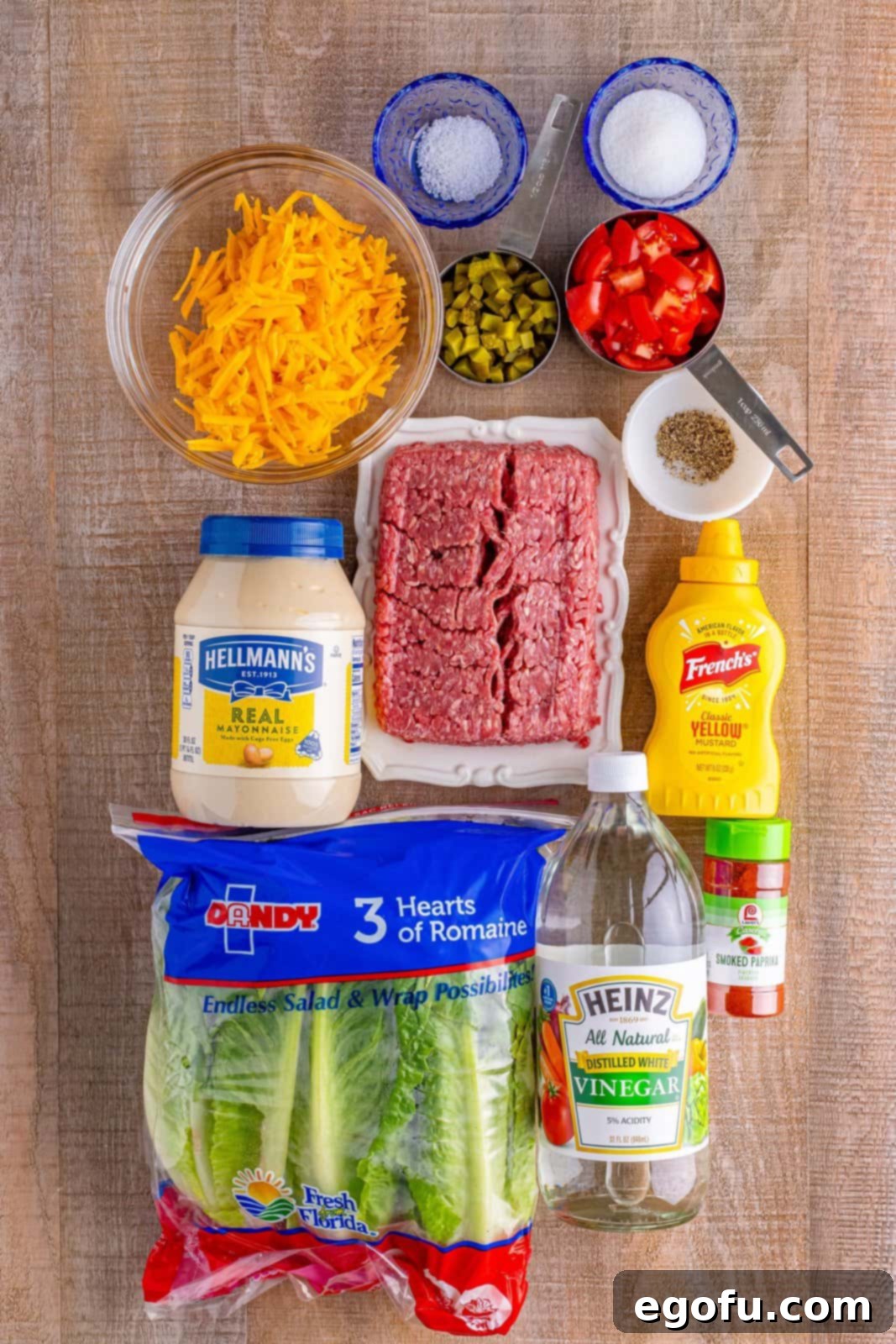 A flat lay of the essential ingredients for Big Mac Salad, including raw ground beef, a jar of mayonnaise, yellow mustard, a bottle of white vinegar, a spice jar of smoked paprika, a bowl of diced dill pickles, a head of fresh romaine lettuce, and vibrant diced tomatoes. All ingredients are neatly arranged, ready for preparation.