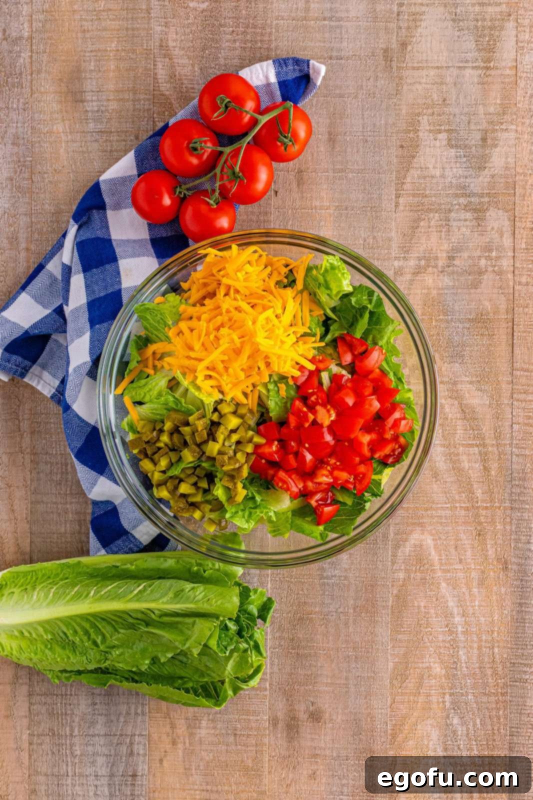 A large clear glass bowl containing a fresh mix of Big Mac salad ingredients, including crisp chopped lettuce, vibrant diced tomatoes, and shredded cheddar cheese, waiting for the protein and dressing.
