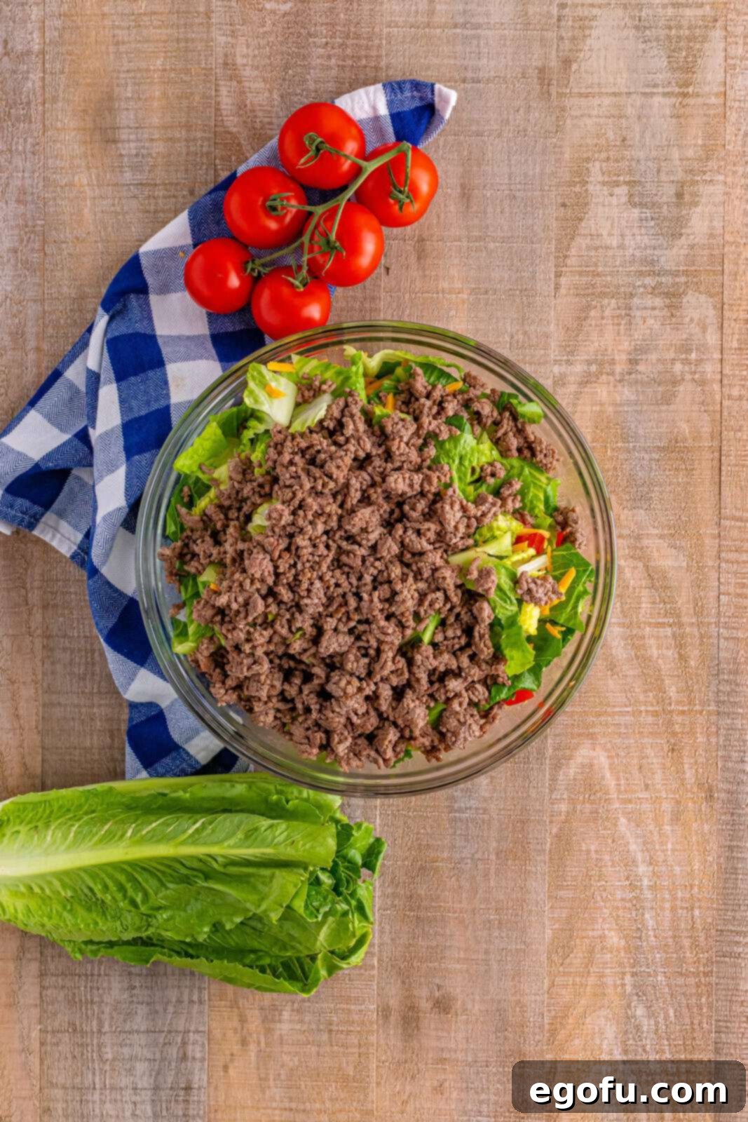 Cooked and seasoned ground beef being added to the large glass bowl of salad ingredients, ready to be mixed together for the Big Mac Salad. The beef adds a substantial and savory element to the fresh greens.