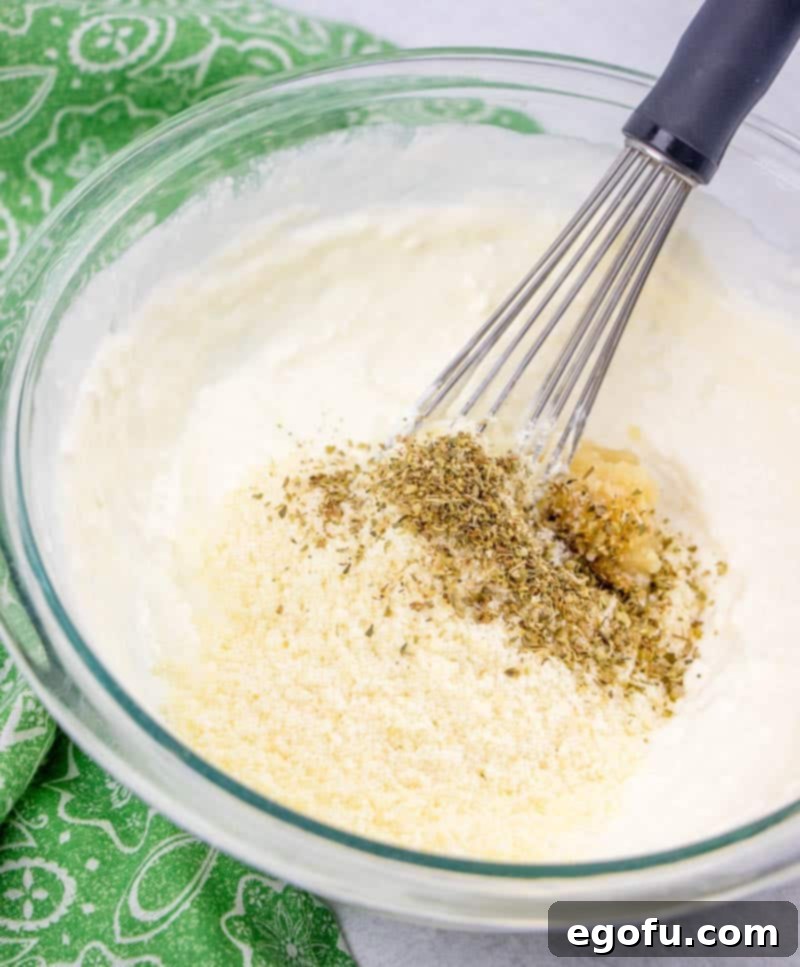 Cream cheese, heavy cream, oregano, Parmesan cheese, and minced garlic whisked together in a glass bowl, forming a smooth and rich sauce.