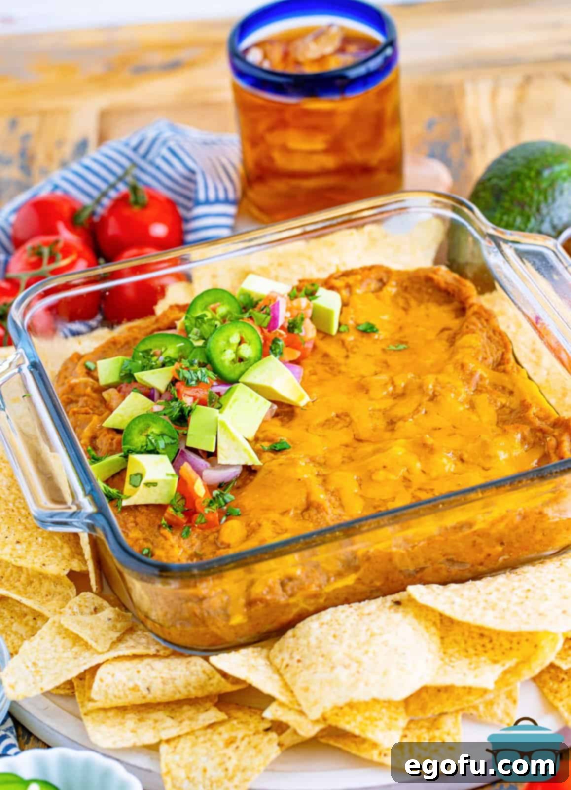 fully baked refried bean dip with toppings shown with tortilla chips and a glass of iced tea in the background. 