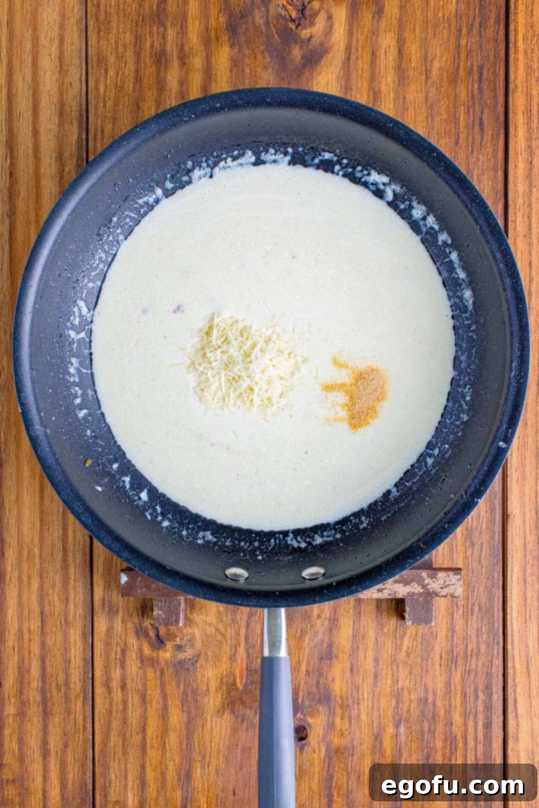 Garlic powder and more Parmesan cheese being stirred into the cream mixture in a saucepan.