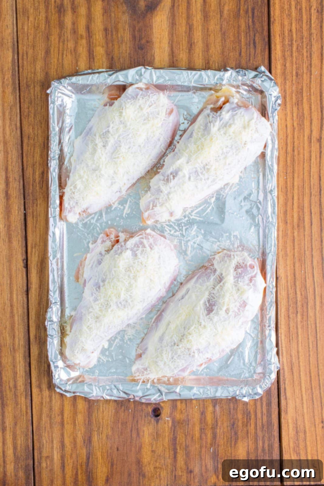 Raw chicken breasts, now coated with mayonnaise, with Parmesan cheese generously sprinkled on top of each.