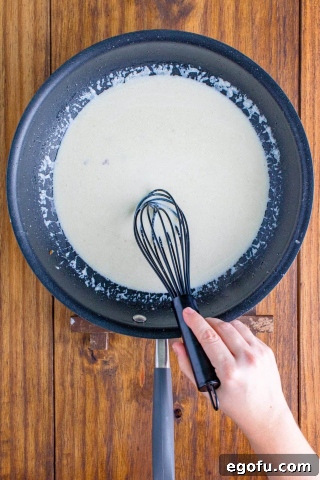 Heavy cream being whisked into melted bacon fat in a saucepan, creating a smooth mixture.