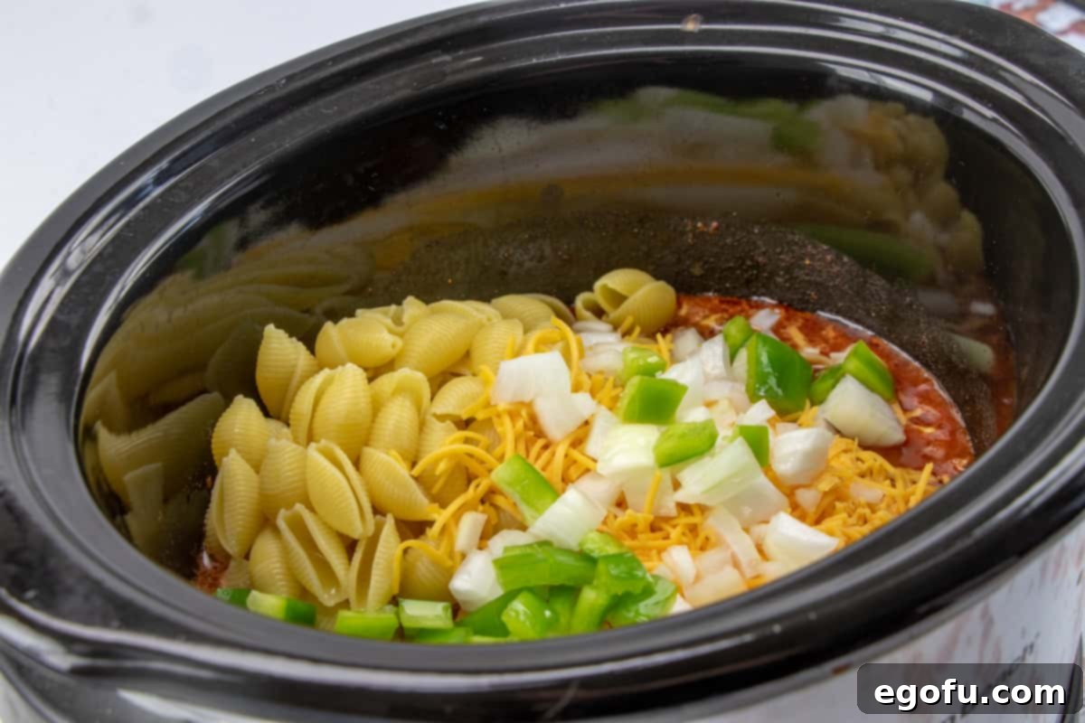 Pasta shells, green pepper, and onions added to ground beef, diced tomatoes, and taco seasoning in an oval slow cooker.