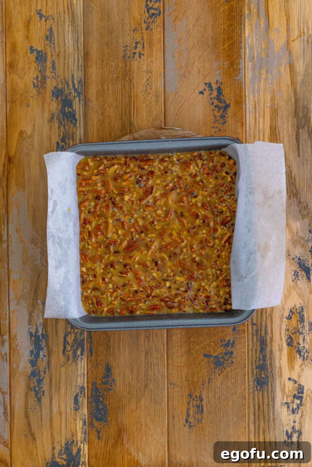 The raw pecan pie filling mixture being poured over the par-baked shortbread crust in a square baking dish, ready for its final bake.