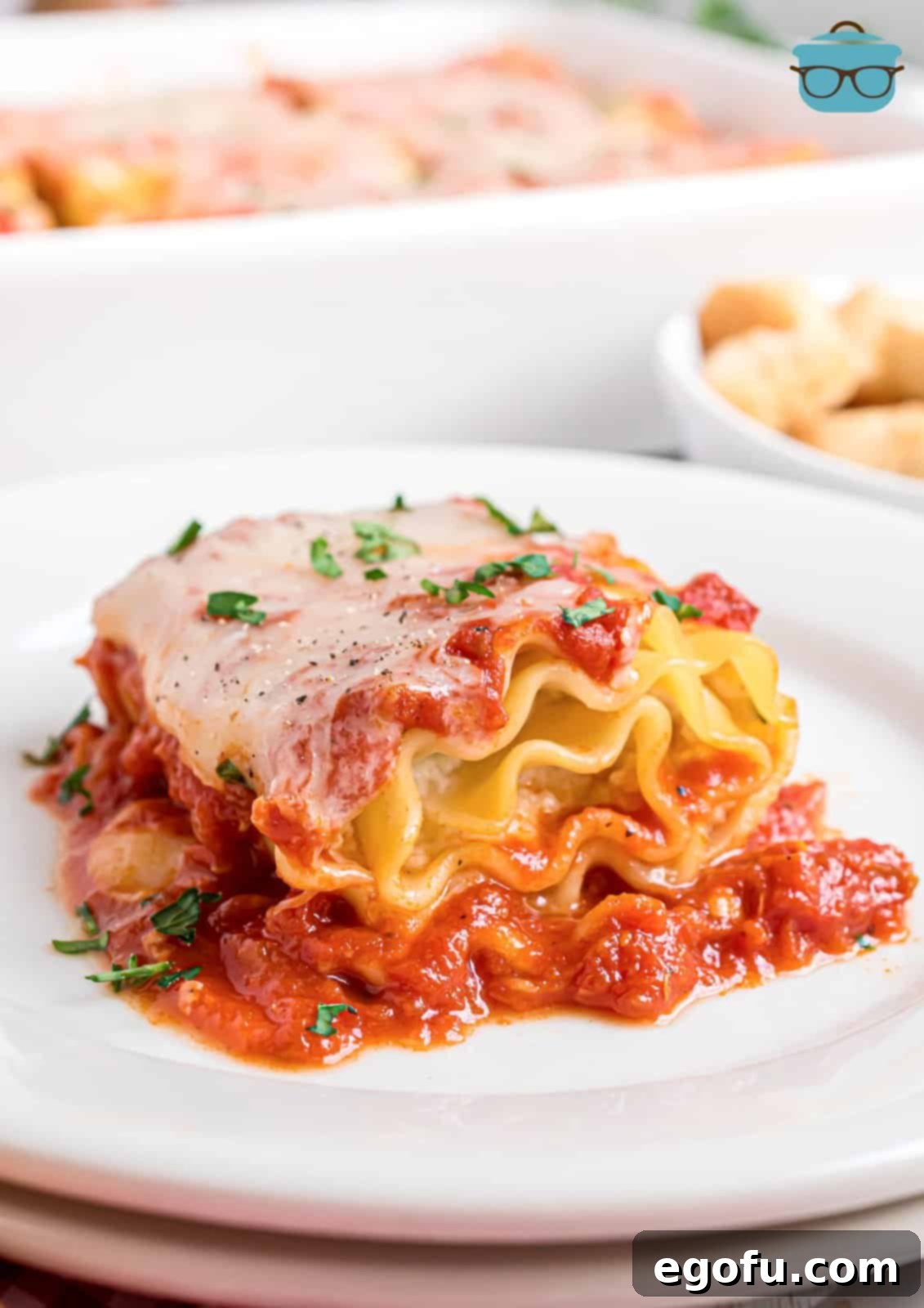 A beautifully presented lasagna roll up on a stack of white plates, with a baking dish of more roll ups blurred in the background, hinting at a delicious, ready-to-serve meal.