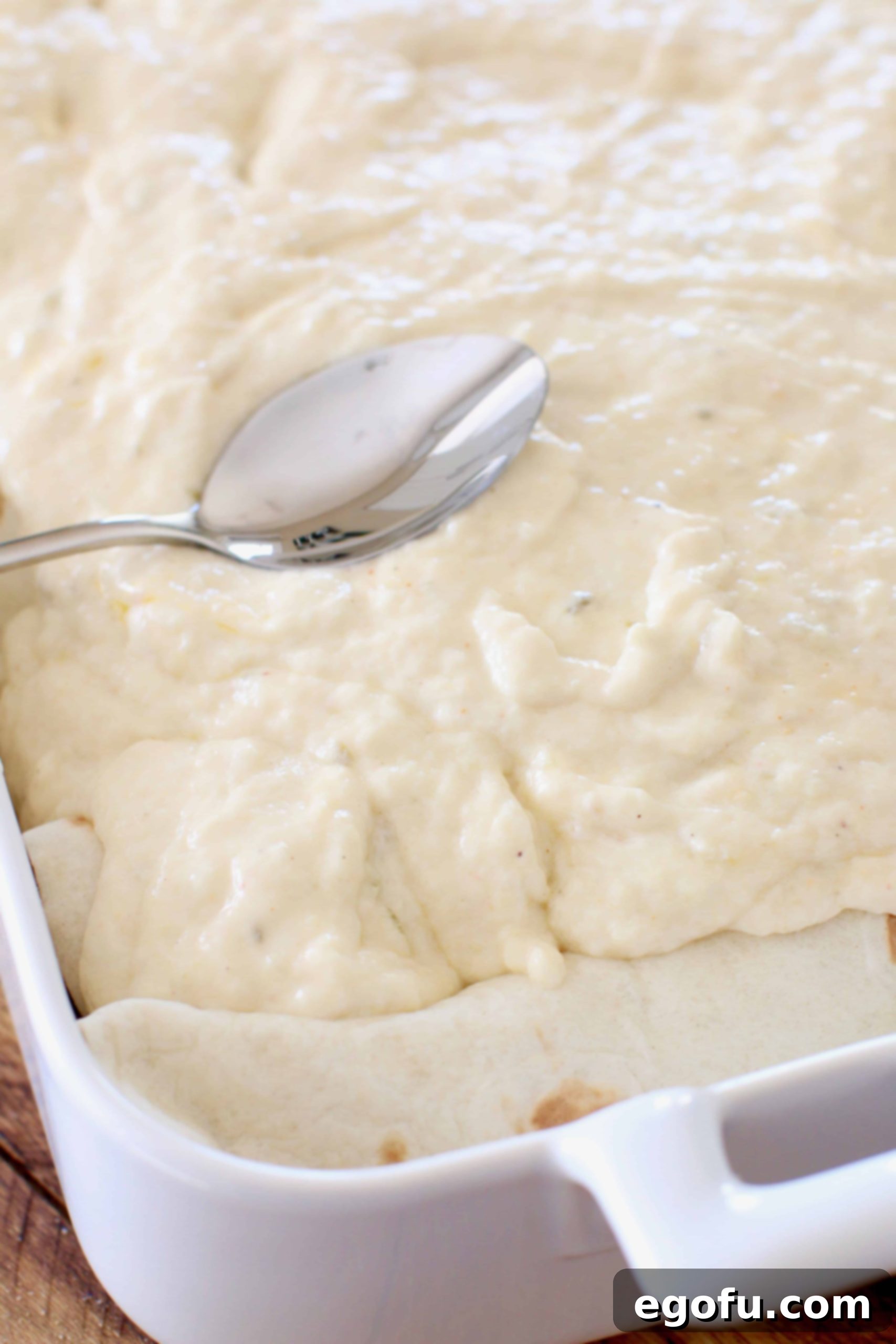 creamy cheese sauce on top of shrimp enchiladas being spread out with the back of a silver spoon.