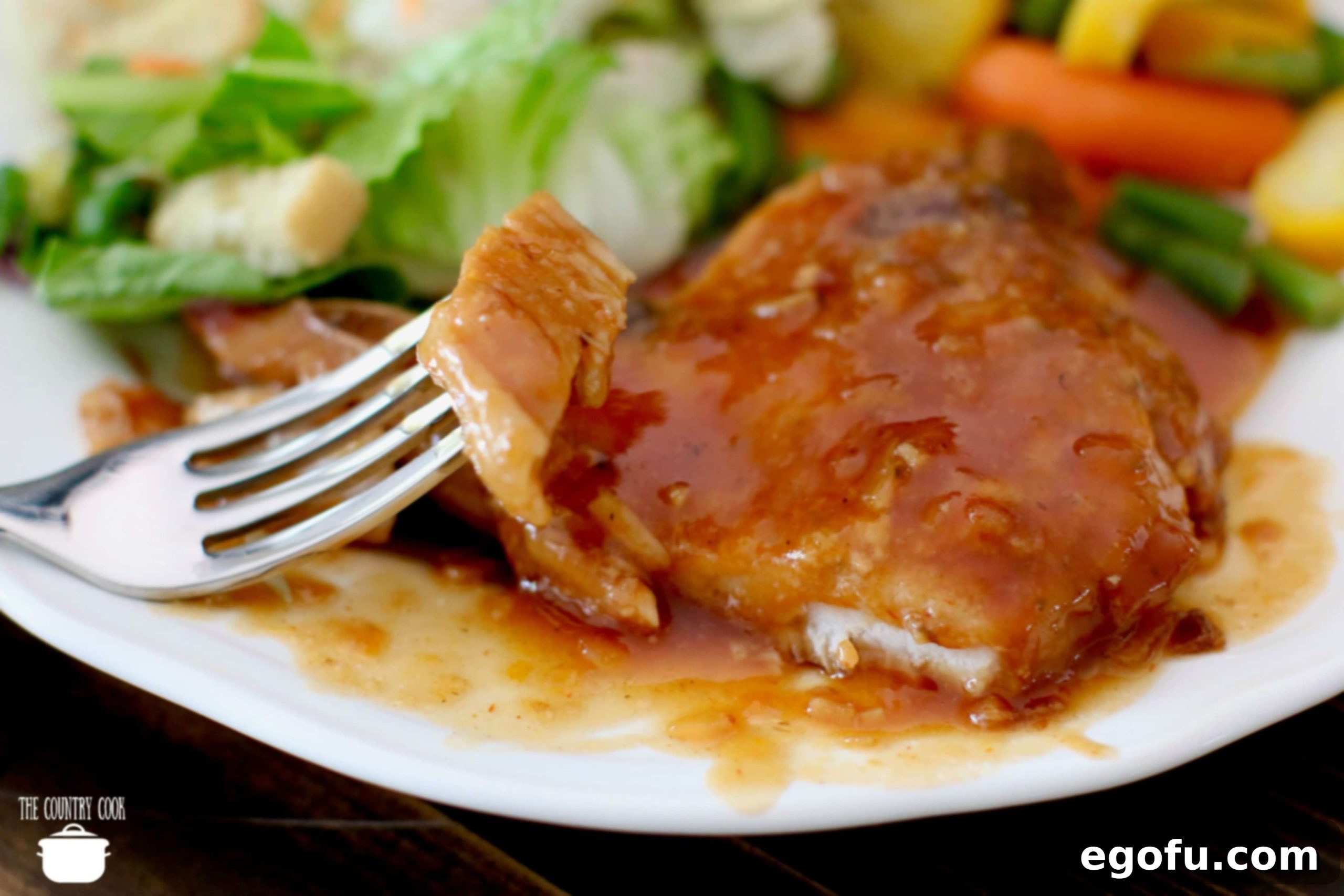 Fork shown holding up a slice of BBQ pork chops on a white plate with a salad and vegetables in the background.